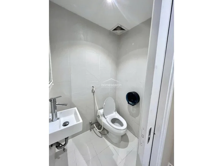 This compact bathroom is finished with textured, light-colored wall tiles and a polished white floor. It is equipped with a modern vessel sink set on a black vanity top, a wall-mounted toilet, and a bidet sprayer, reflecting a well-maintained, contemporary design.