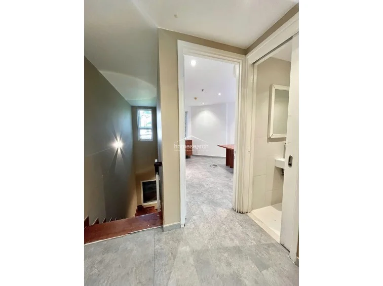 This transitional space connects a staircase area with a private room, featuring a neutral beige color scheme and consistent grey floor tiles. To the right, a compact bathroom with a sliding door is visible, showcasing a wall-mounted sink and a clean, minimalist design.