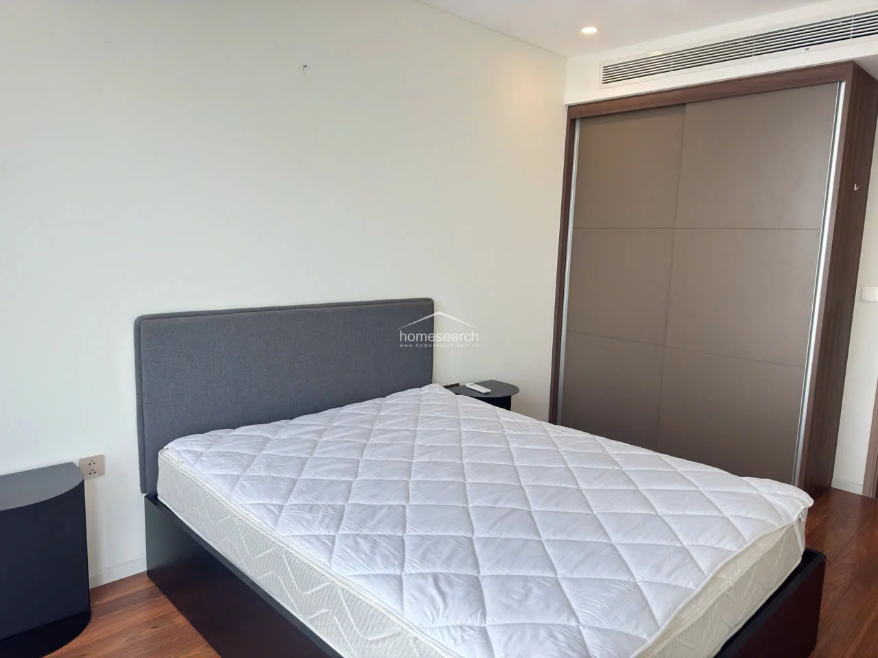 This bedroom features a clean, minimalist design with warm wood flooring and neutral white walls. It includes a built-in floor-to-ceiling wardrobe with a sleek, dark wood finish and a modern integrated air conditioning unit above.