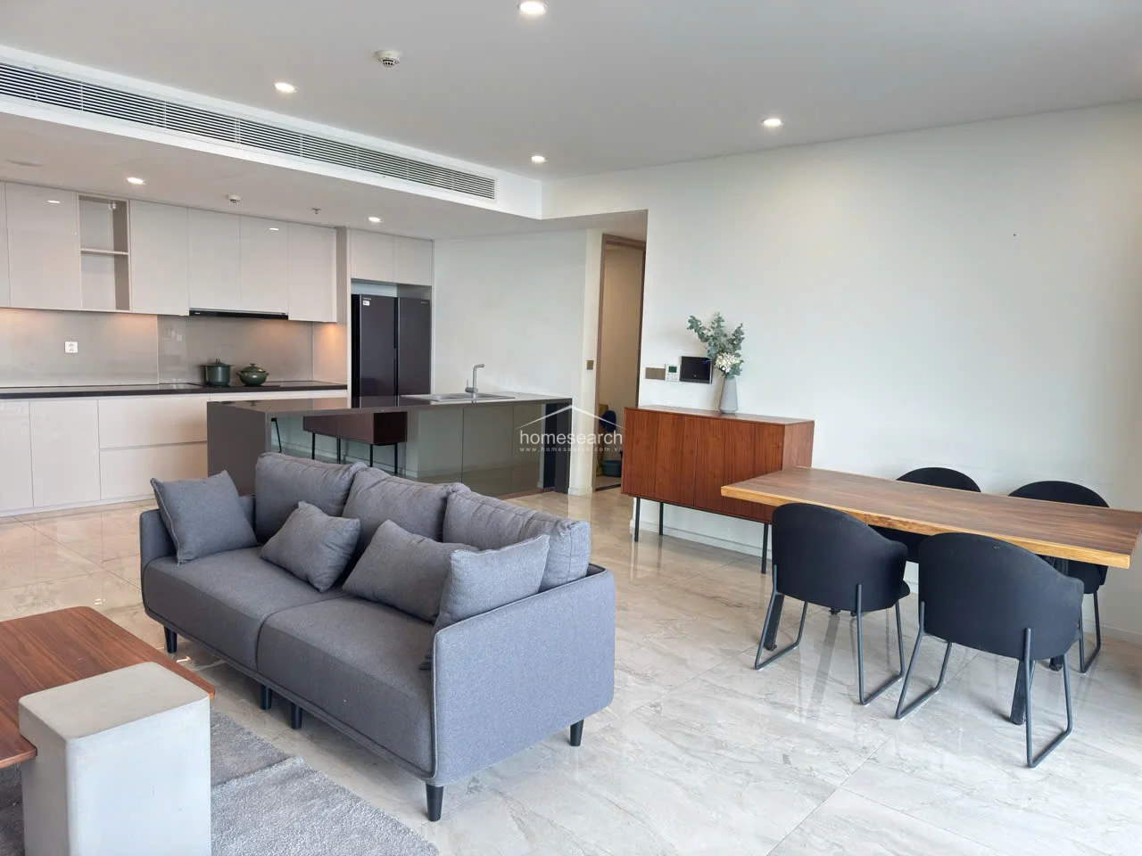 This open-concept living and dining area features a modern, minimalist design with high-gloss cabinetry and a sleek kitchen island. The space is anchored by polished marble-style flooring and neutral wall finishes, complemented by recessed lighting and a central air conditioning vent. The layout flows seamlessly from the kitchen into a comfortable lounge area, creating an ideal space for entertaining.