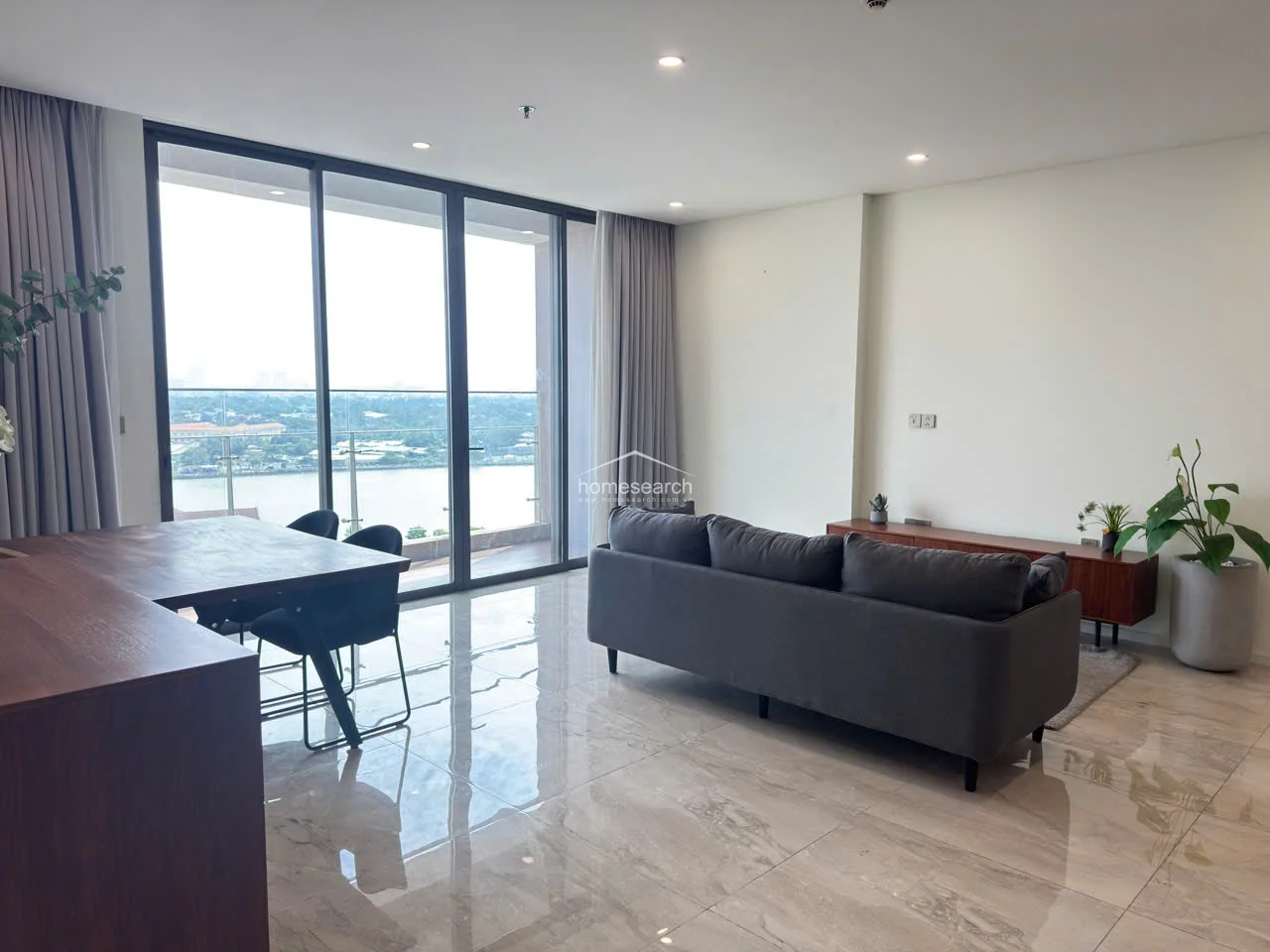The living room is bathed in natural light provided by floor-to-ceiling glass sliding doors that open onto a balcony with expansive river views. The room is furnished with a contemporary grey sofa and a wooden media console, maintaining a clean, uncluttered aesthetic. The neutral color palette and high-quality finishes suggest a well-maintained, modern property.