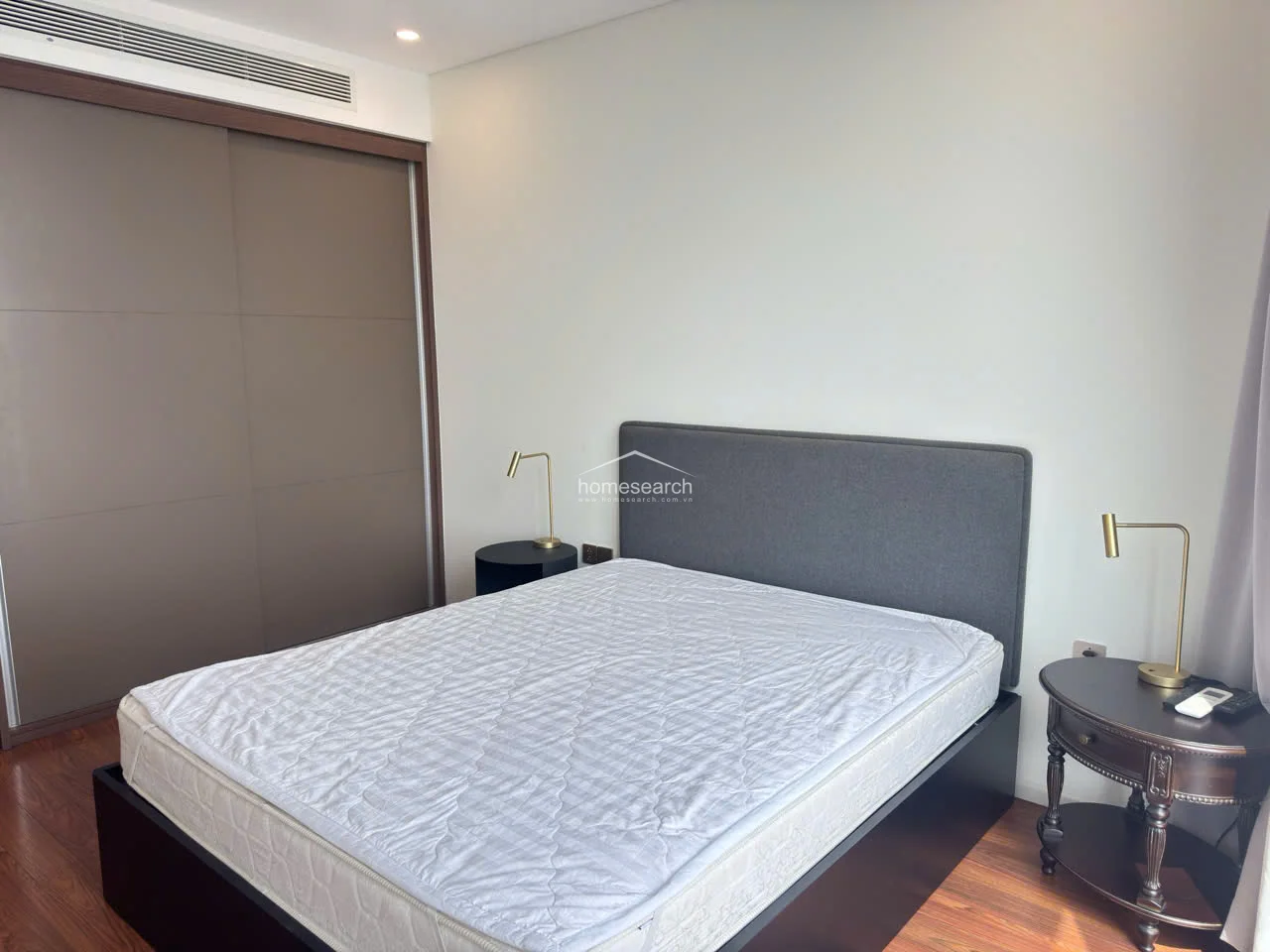This angle highlights the bedroom's cohesive aesthetic, featuring a grey upholstered headboard and matching dark wood bedside tables with elegant gold-accented lamps. The space is well-maintained and provides a functional, contemporary sleeping environment.