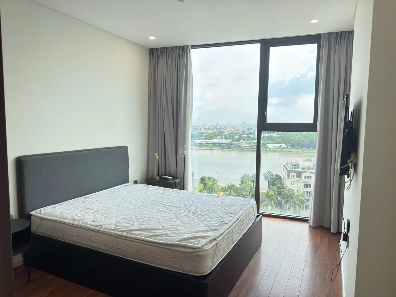 This bedroom offers a stunning, unobstructed river view through floor-to-ceiling windows draped in soft grey curtains. The room maintains a consistent design language with rich wood flooring and a minimalist layout that maximizes the impact of the natural light and scenery.