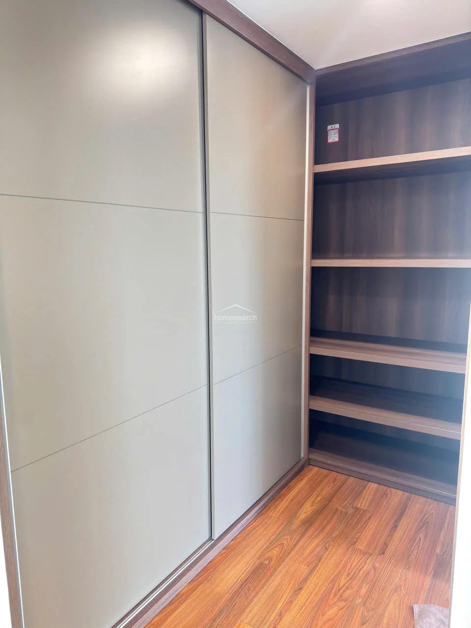This walk-in closet space is designed with a modern aesthetic, featuring sleek grey sliding doors and open shelving in a rich wood finish. The warm wood flooring adds a touch of elegance, creating a well-organized and stylish storage solution.