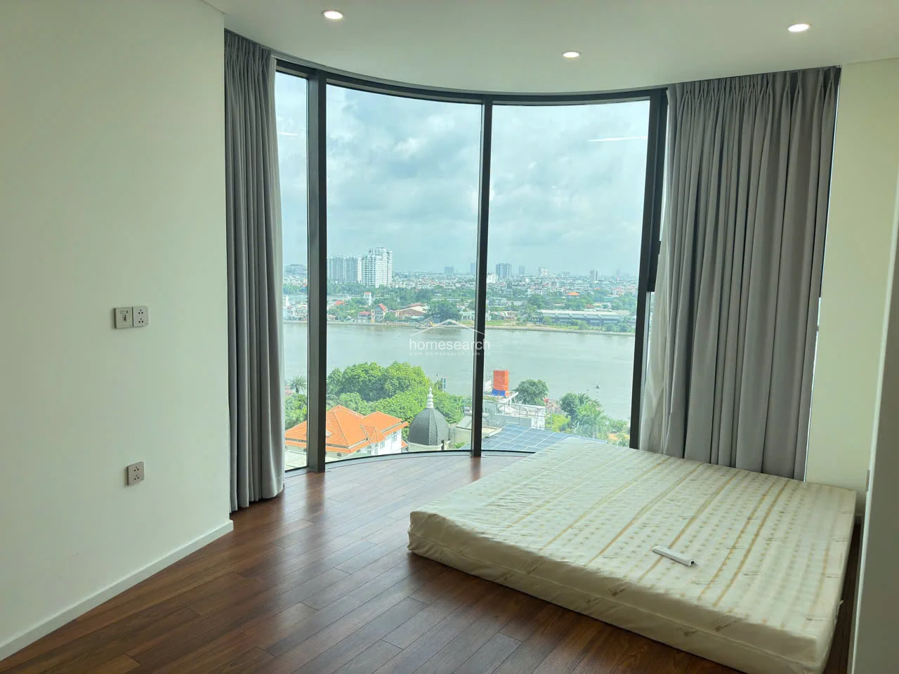 This image showcases a unique curved window design that creates an expansive, panoramic view of the river and cityscape. The room is currently sparsely furnished with a low-profile mattress, emphasizing the architectural interest of the curved glass walls and the bright, airy atmosphere.