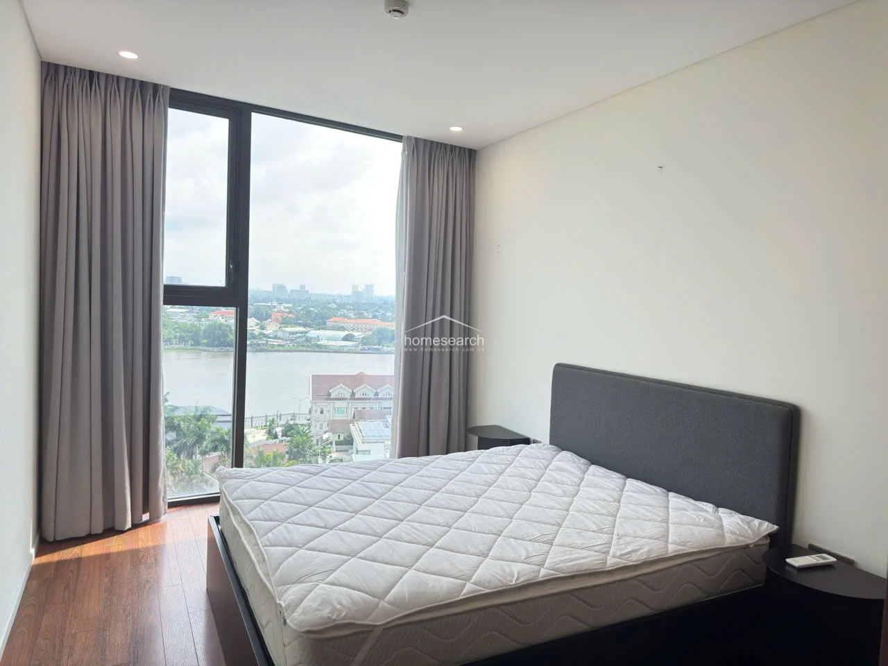 The bedroom maintains a minimalist and tranquil atmosphere, featuring warm wood-look flooring and a simple grey upholstered bed frame. A large floor-to-ceiling window captures the same impressive river view, allowing for plenty of natural light. The room is kept clean and uncluttered, emphasizing the high-quality construction and modern design of the residence.