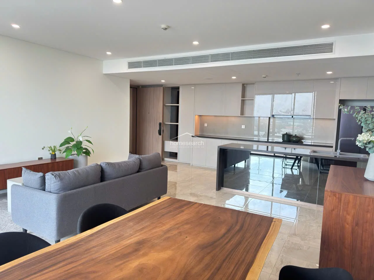 This view highlights the functional kitchen layout, which includes an integrated refrigerator and ample cabinet storage. The dining area is defined by a natural wood-edge table and dark chairs, providing a warm contrast to the cool tones of the marble flooring. The entrance area includes built-in shelving, demonstrating efficient use of space in this modern apartment.