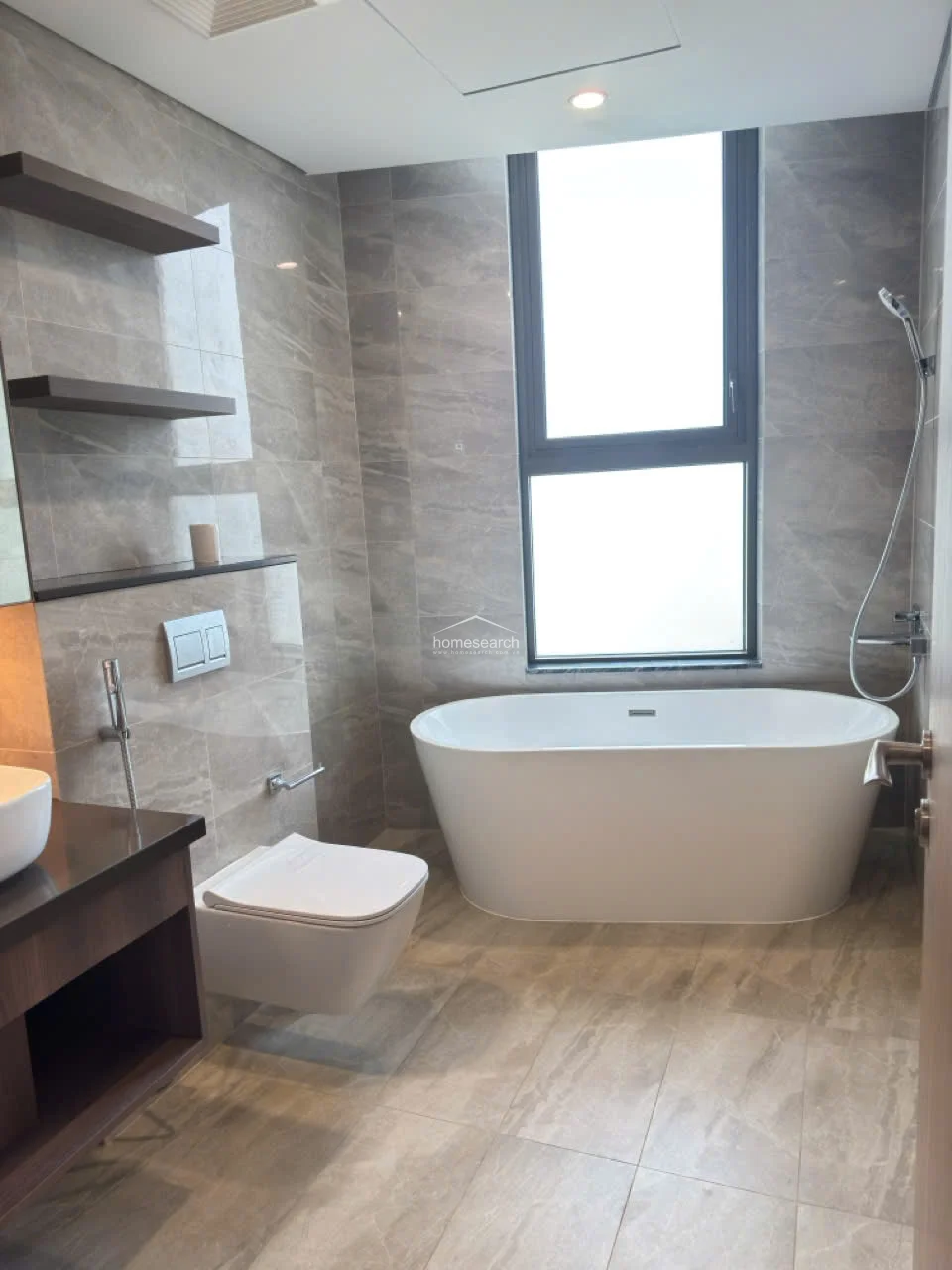 This luxurious bathroom is finished in sophisticated grey marble-effect tiling and features a contemporary freestanding soaking tub positioned beneath a large window for natural light. The wall-mounted toilet and floating vanity contribute to a clean, minimalist flow, while built-in shelving provides convenient storage.