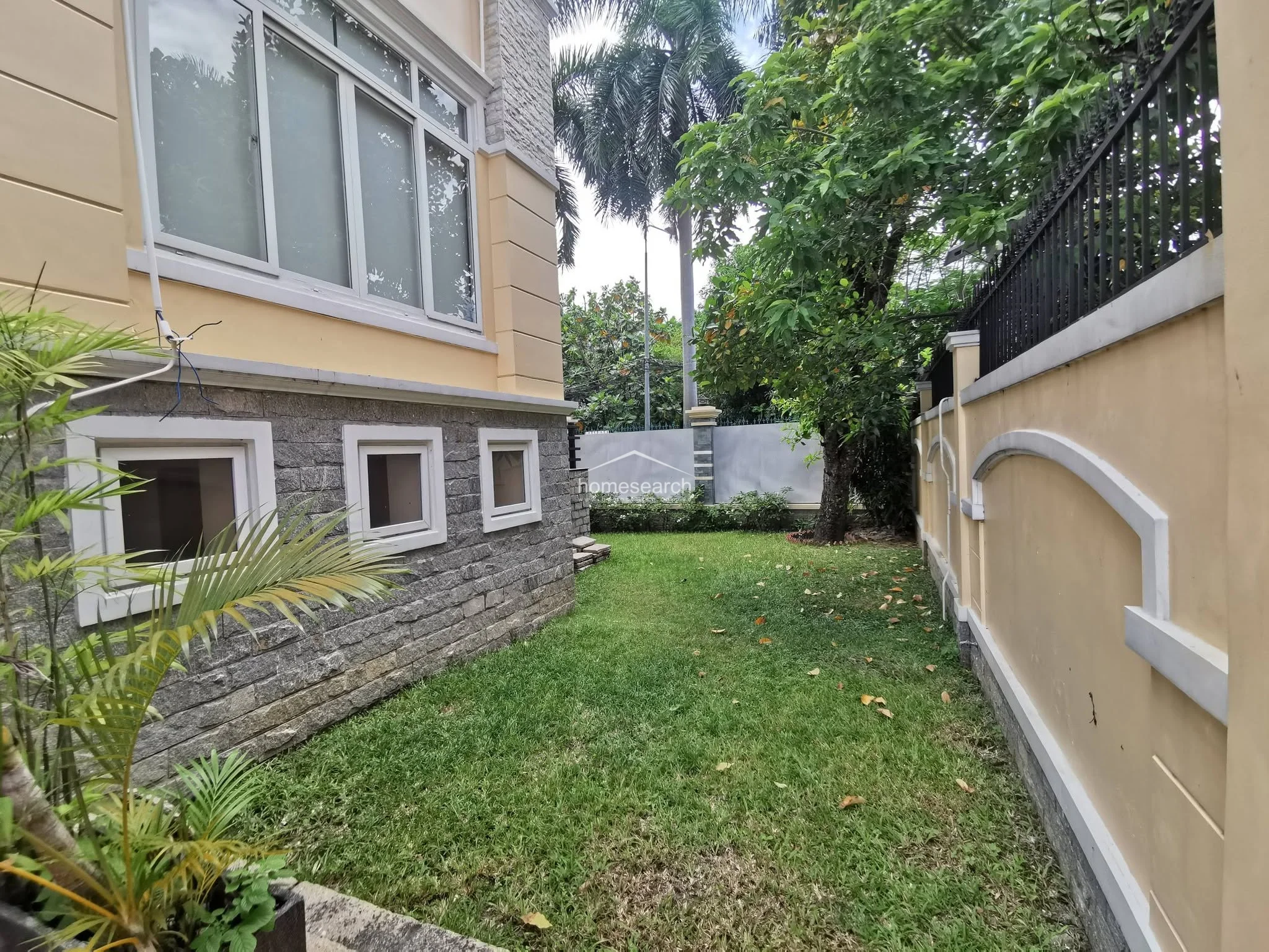 This side yard offers a quiet, grassy lawn bordered by a high perimeter wall and mature trees, providing excellent privacy. The house exterior features a combination of light-colored stucco and rustic stone cladding, with multiple windows allowing for natural light.