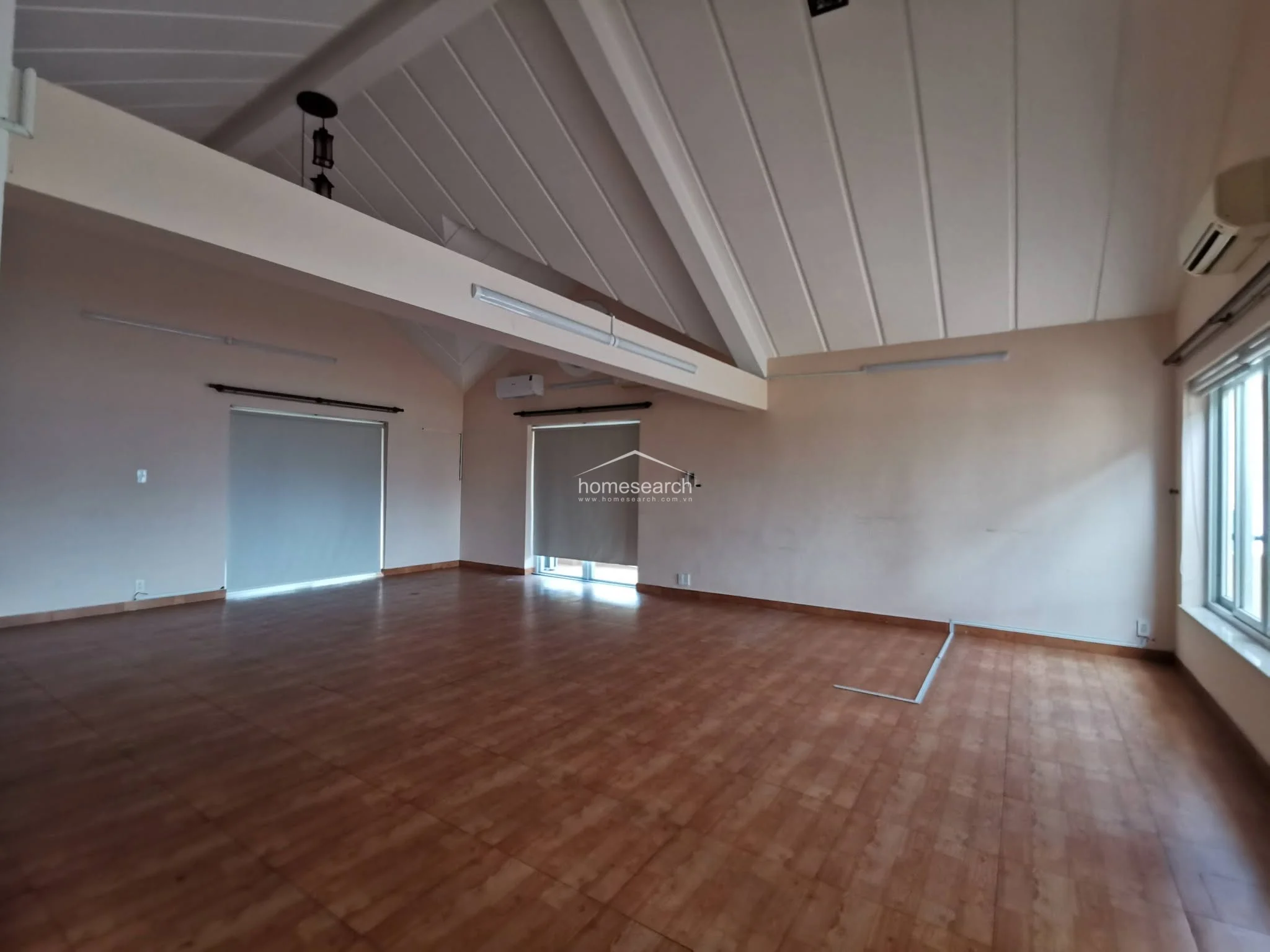 This expansive room features a vaulted ceiling with exposed beams, providing a sense of height and openness. It is equipped with a wall-mounted air conditioning unit and large windows with roller blinds, while the warm-toned wood-look flooring adds a cozy, inviting atmosphere to the versatile space.