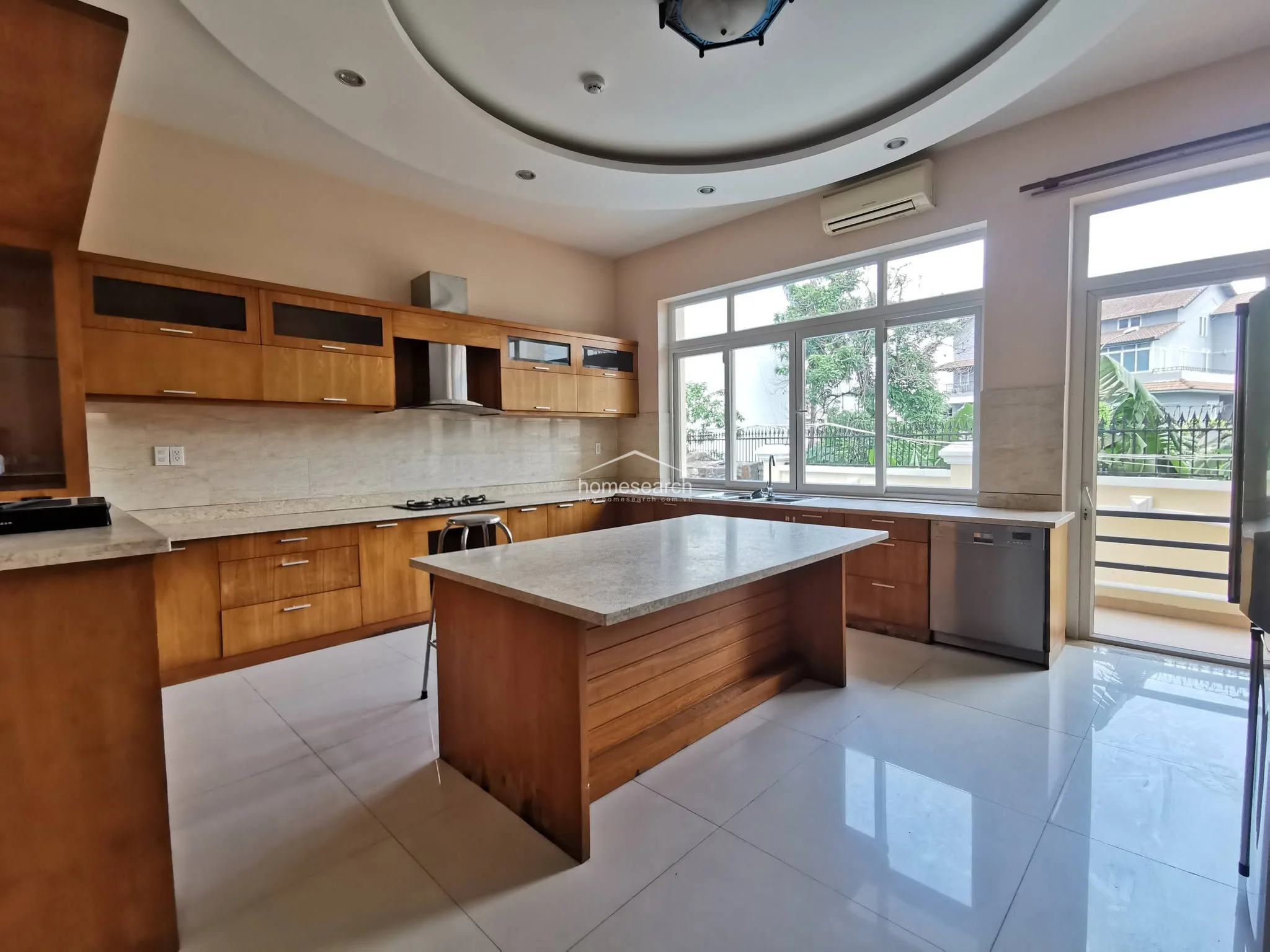 This expansive kitchen features warm wood cabinetry, a large central island with a stone countertop, and glossy white floor tiles. It is equipped with a built-in dishwasher, a stainless steel range hood, and a ceiling fan, all set under a sophisticated circular recessed ceiling design.