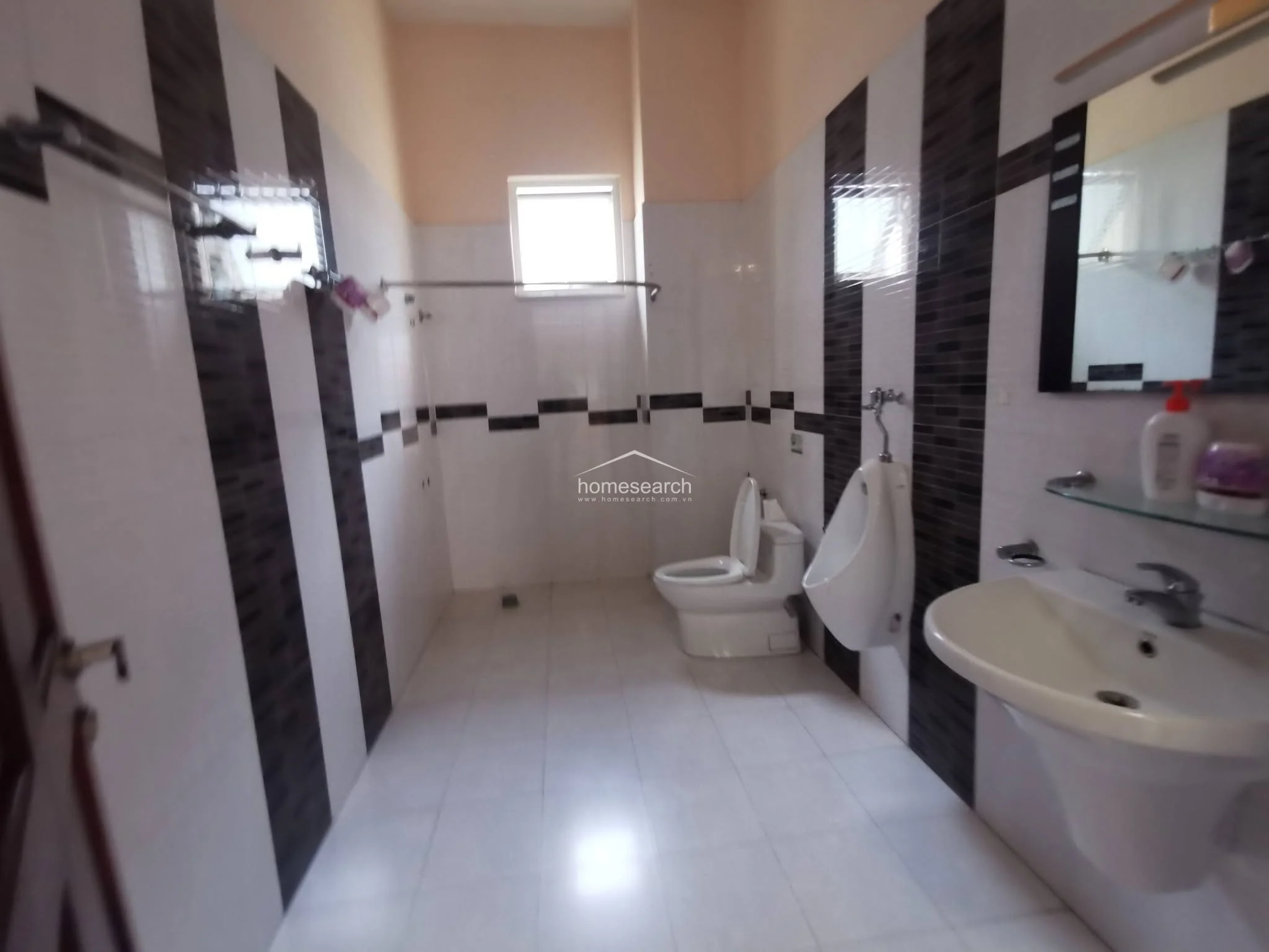 This modern bathroom is characterized by a bold, high-contrast design featuring vertical black and white tiled stripes on the walls. The layout includes a standard toilet, a wall-mounted urinal, and a pedestal sink, all maintained in clean condition with a small window providing natural ventilation.