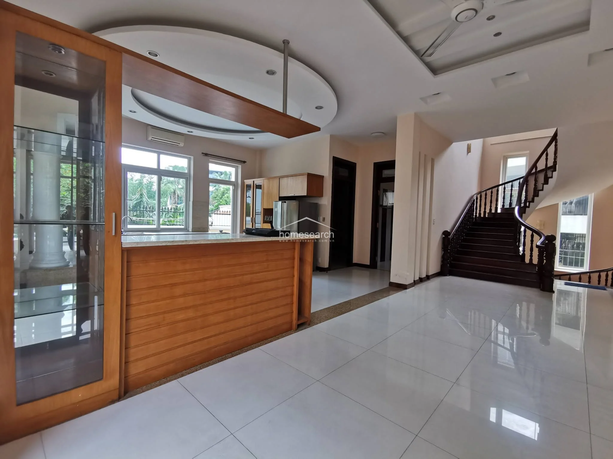 This open-concept living and kitchen area flows seamlessly into a grand staircase with a dark wood banister and decorative spindles. The space is brightened by large windows and features a built-in glass display cabinet, high ceilings with recessed lighting, and polished white tile flooring.