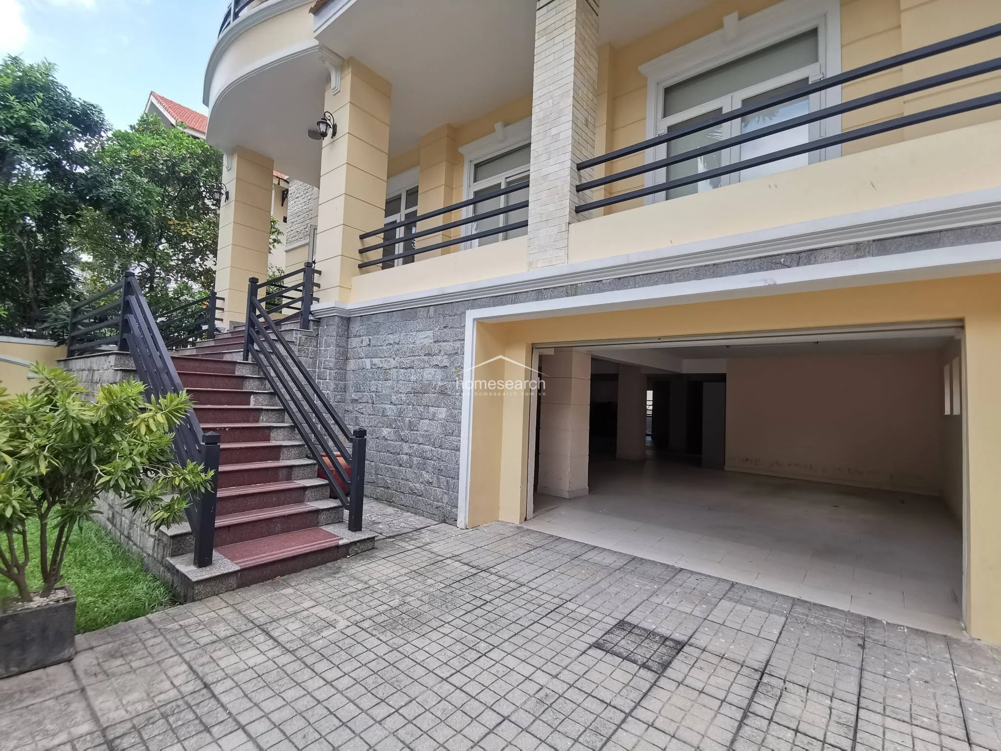 This exterior view shows a multi-level home with a spacious, open-air garage on the ground floor and a balcony above. A grand staircase with dark wood railings leads up to the main living level, surrounded by mature greenery and paved walkways.