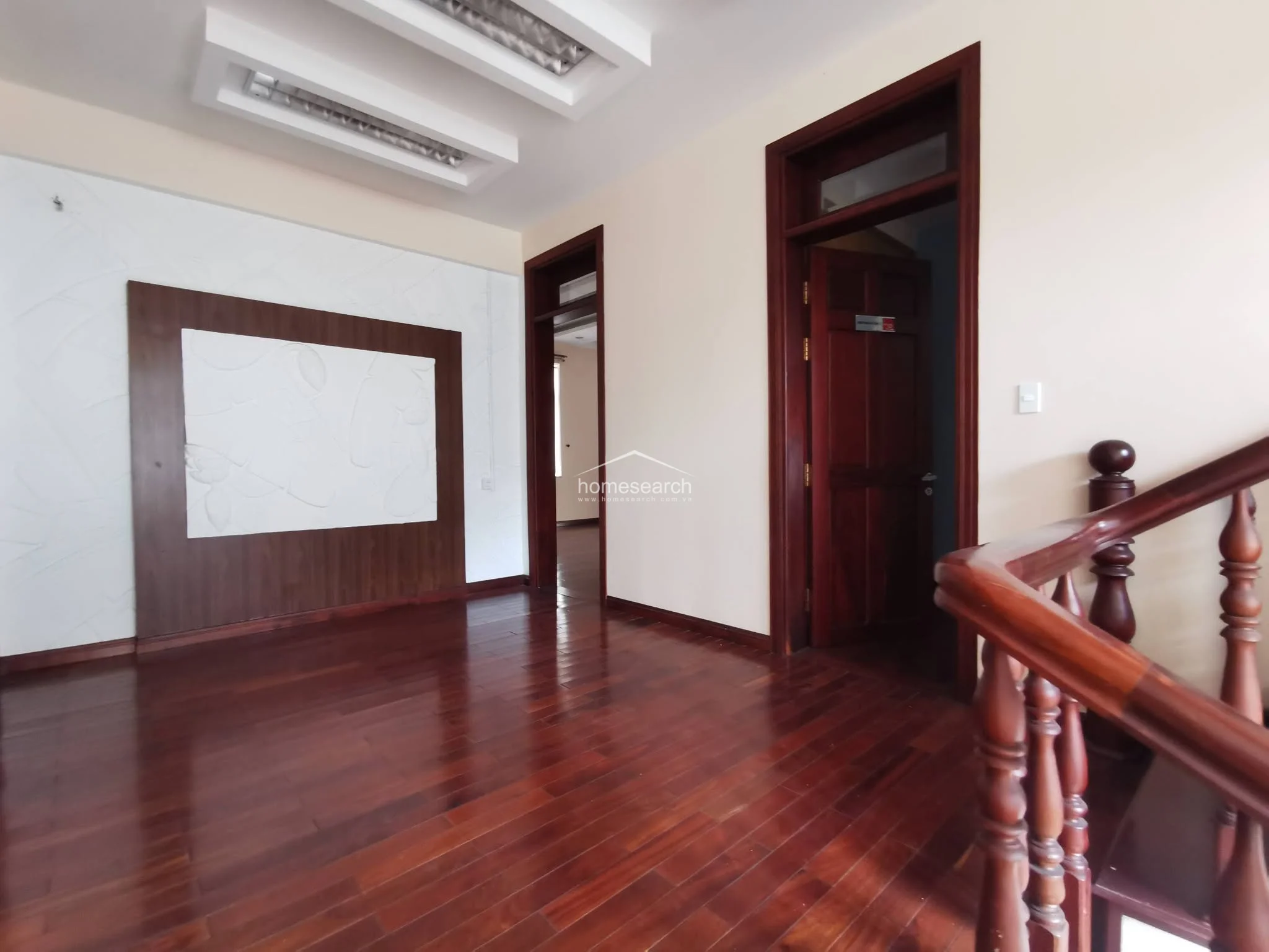 This upper-level landing or hallway showcases rich, polished dark wood flooring and a classic wooden staircase with an ornate balustrade. The walls are painted in a neutral cream tone, complemented by a decorative wall panel feature and traditional dark wood door frames that lead into private rooms.