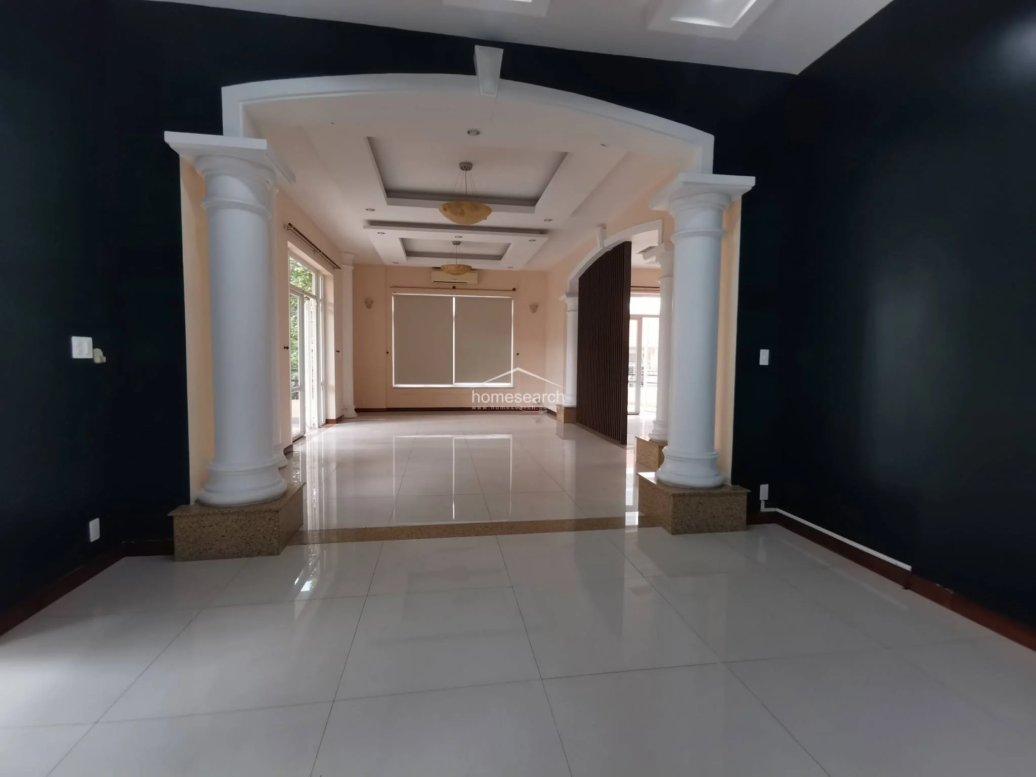 This grand living area features a dramatic open-plan layout defined by two prominent white columns and a sophisticated coffered ceiling with integrated lighting. The space is finished with high-gloss white tile flooring and bold black accent walls, creating a striking contrast that flows seamlessly into adjacent rooms.