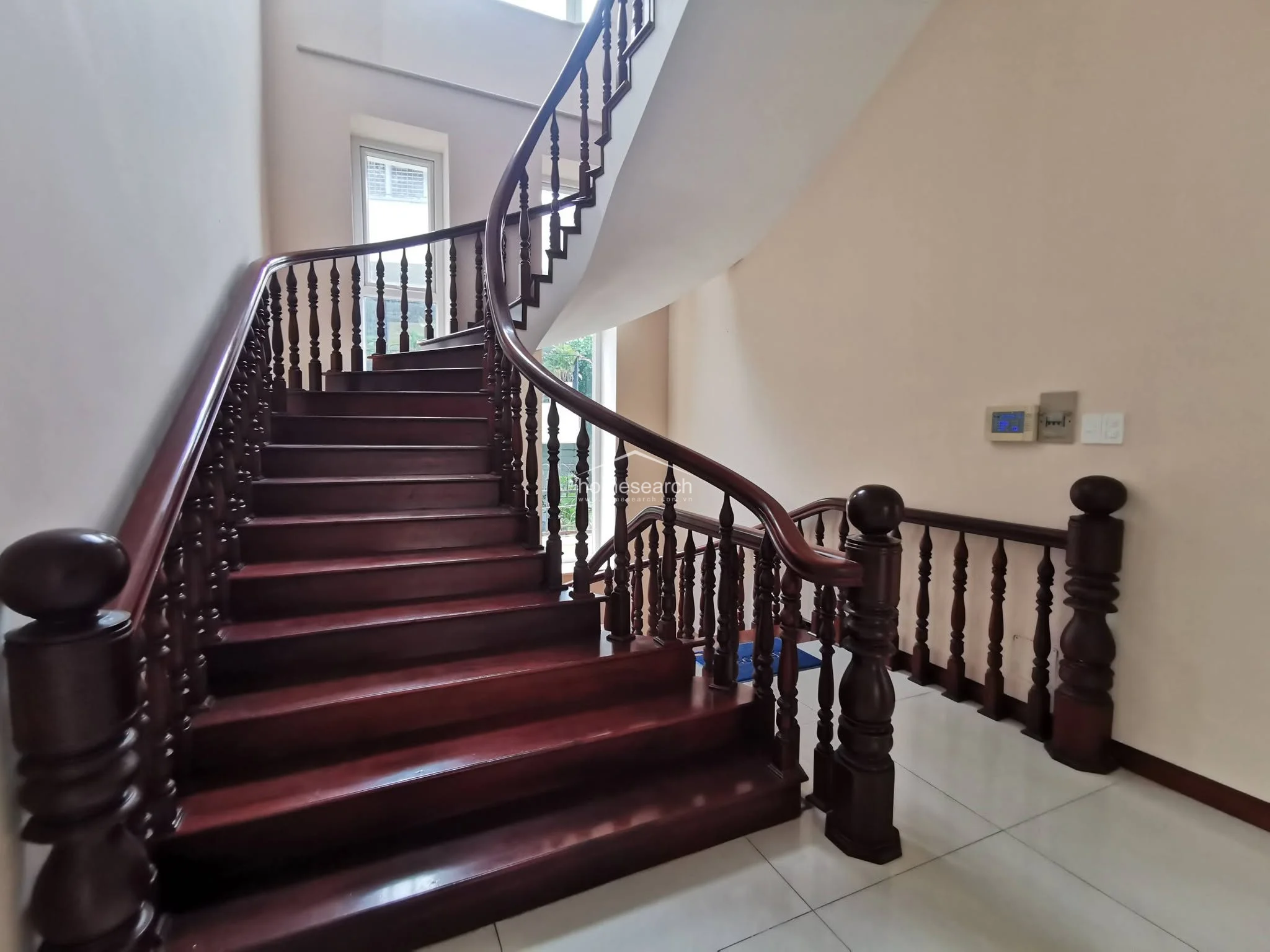 This elegant staircase serves as a central architectural feature, constructed from dark, polished wood with a sweeping curved handrail and matching balusters. The area is brightened by a tall window that invites natural light, while the neutral walls and white tile flooring maintain a clean, well-maintained aesthetic.