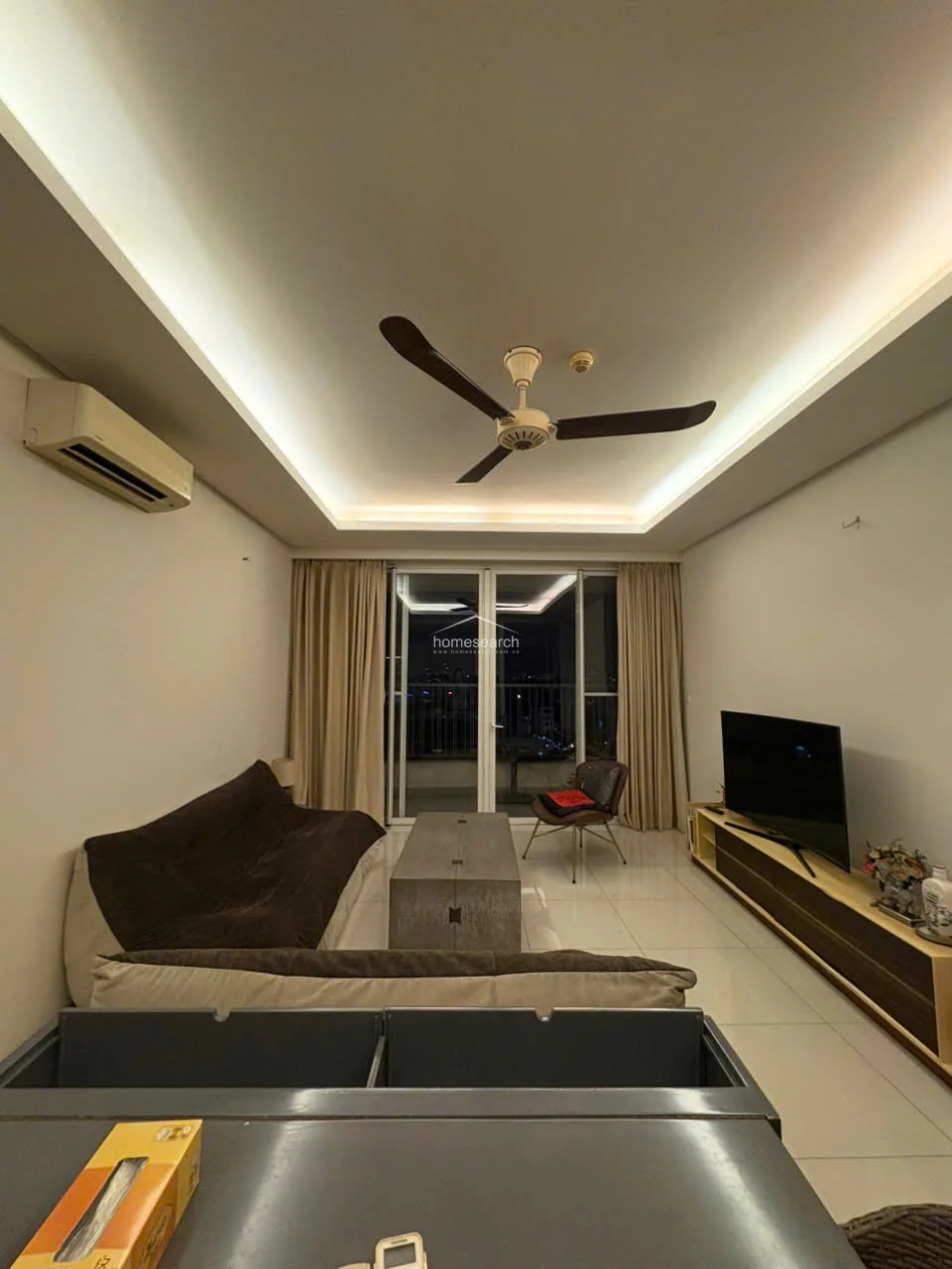 This open-concept living area features a modern tray ceiling with recessed lighting and a sleek ceiling fan, creating an airy and sophisticated atmosphere. The space is finished with polished white tile flooring and connects directly to a balcony through large sliding glass doors that provide ample natural light and city views. The room is equipped with a wall-mounted AC unit and is furnished with a contemporary sofa, a wooden coffee table, and a media console.