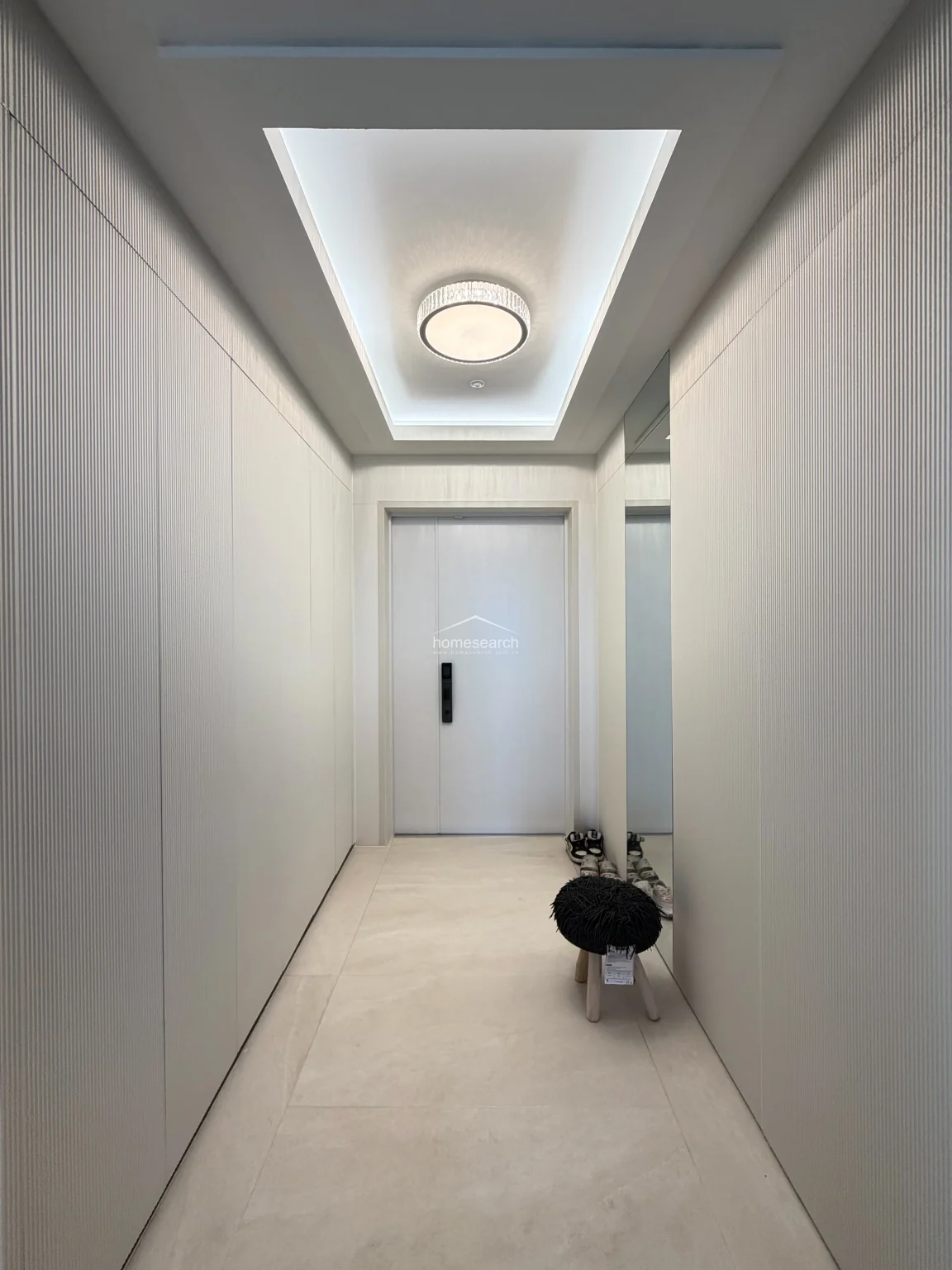 The entryway features a clean, minimalist design with floor-to-ceiling textured wall paneling and a recessed ceiling with soft, ambient lighting. A full-length mirror and a small stool create a welcoming transition space, while the light-toned tile flooring maintains a consistent, airy feel throughout the home.