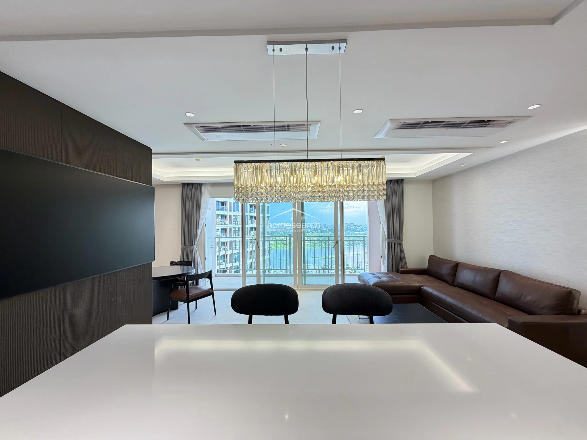 This open-concept living and dining area features a sophisticated, modern aesthetic with a striking crystal chandelier as a focal point. The space is well-lit by large floor-to-ceiling glass doors that provide a scenic view and lead to an outdoor balcony, while the neutral color palette is complemented by rich dark wood accents and high-end finishes.