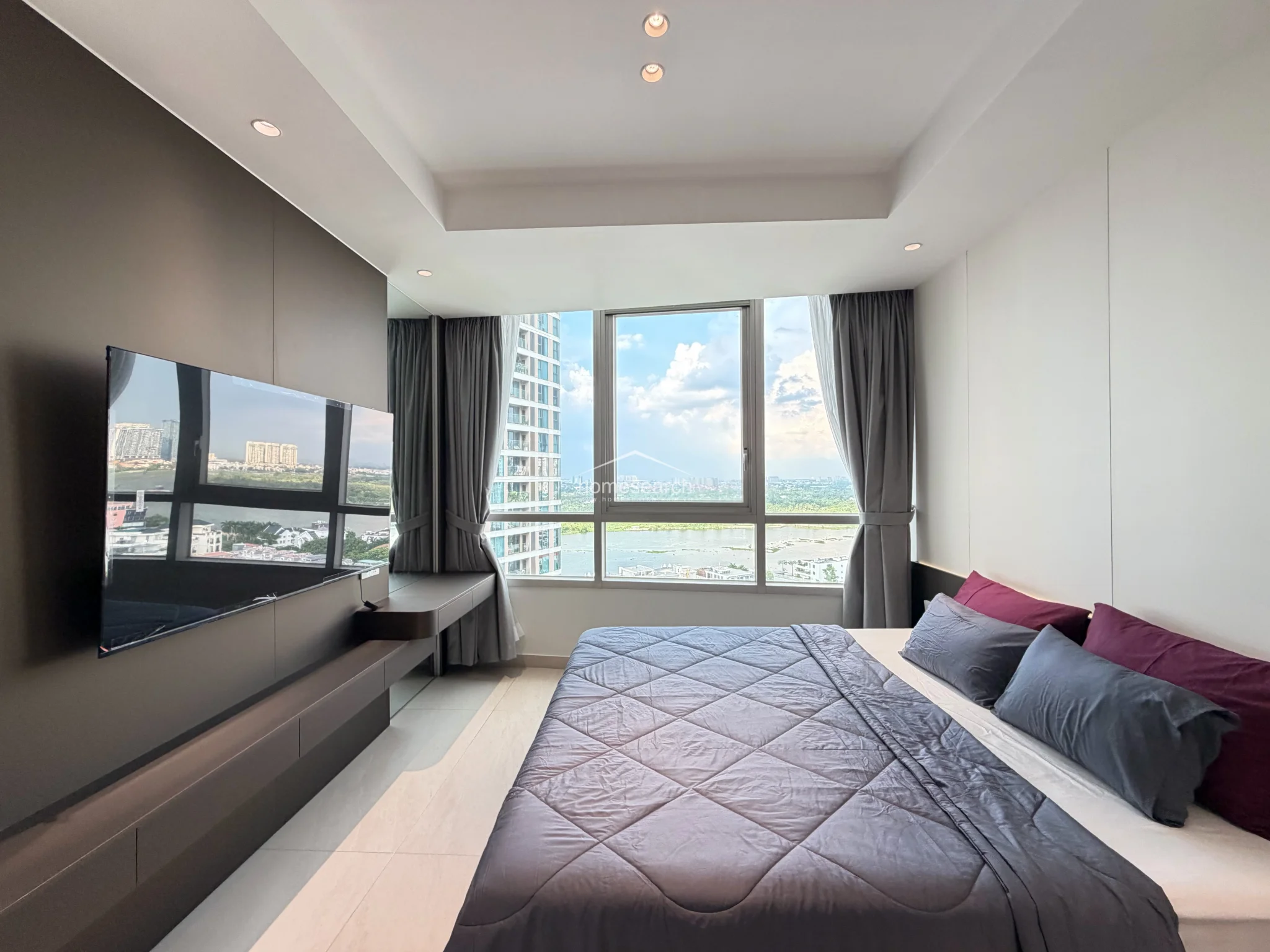 This bedroom is designed with a striking dark-toned accent wall that houses a wall-mounted television and a floating console, creating a sophisticated focal point. The large window is framed by floor-length grey curtains, and the overall condition of the space appears excellent with a clean, contemporary finish.