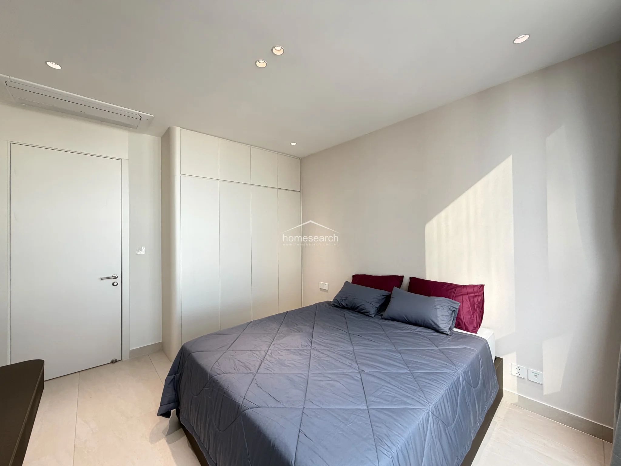 The bedroom layout includes a functional, floor-to-ceiling white wardrobe that provides ample storage while maintaining a minimalist aesthetic. A central ceiling-mounted air conditioning unit ensures climate control, and the room is finished with high-quality, light-toned tile flooring throughout.