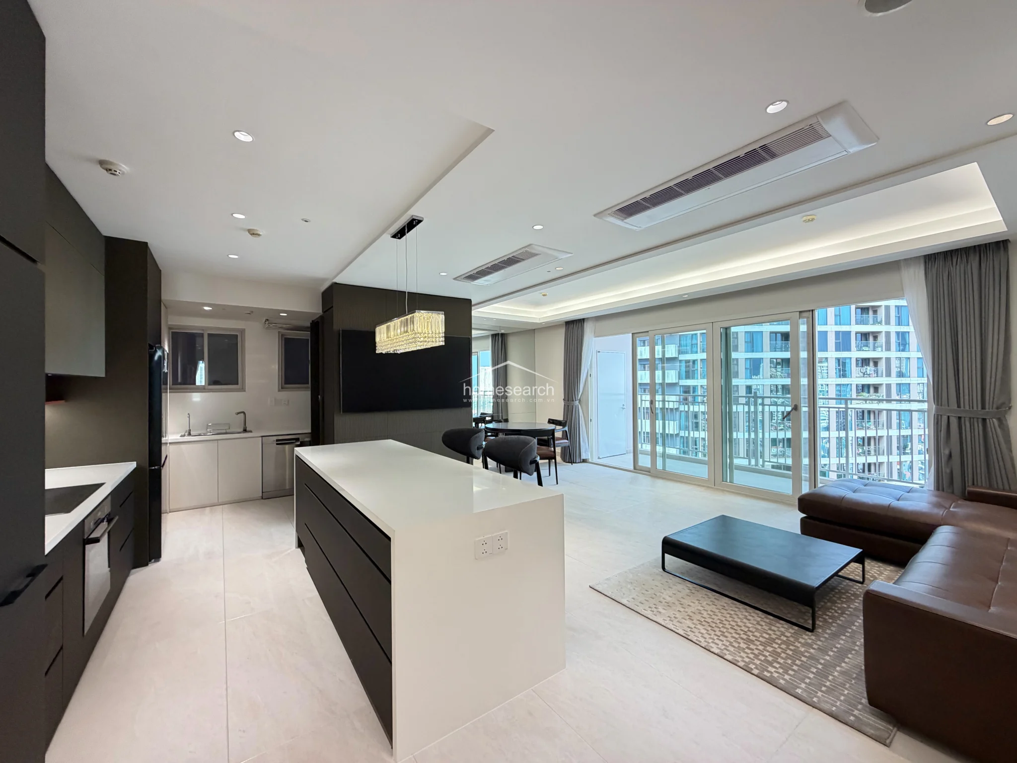 The kitchen is seamlessly integrated into the living space, featuring a sleek white island and dark cabinetry that provides ample storage. It is equipped with modern appliances, including a built-in oven and a large refrigerator, all set against a clean, minimalist backdrop with light-colored tile flooring.