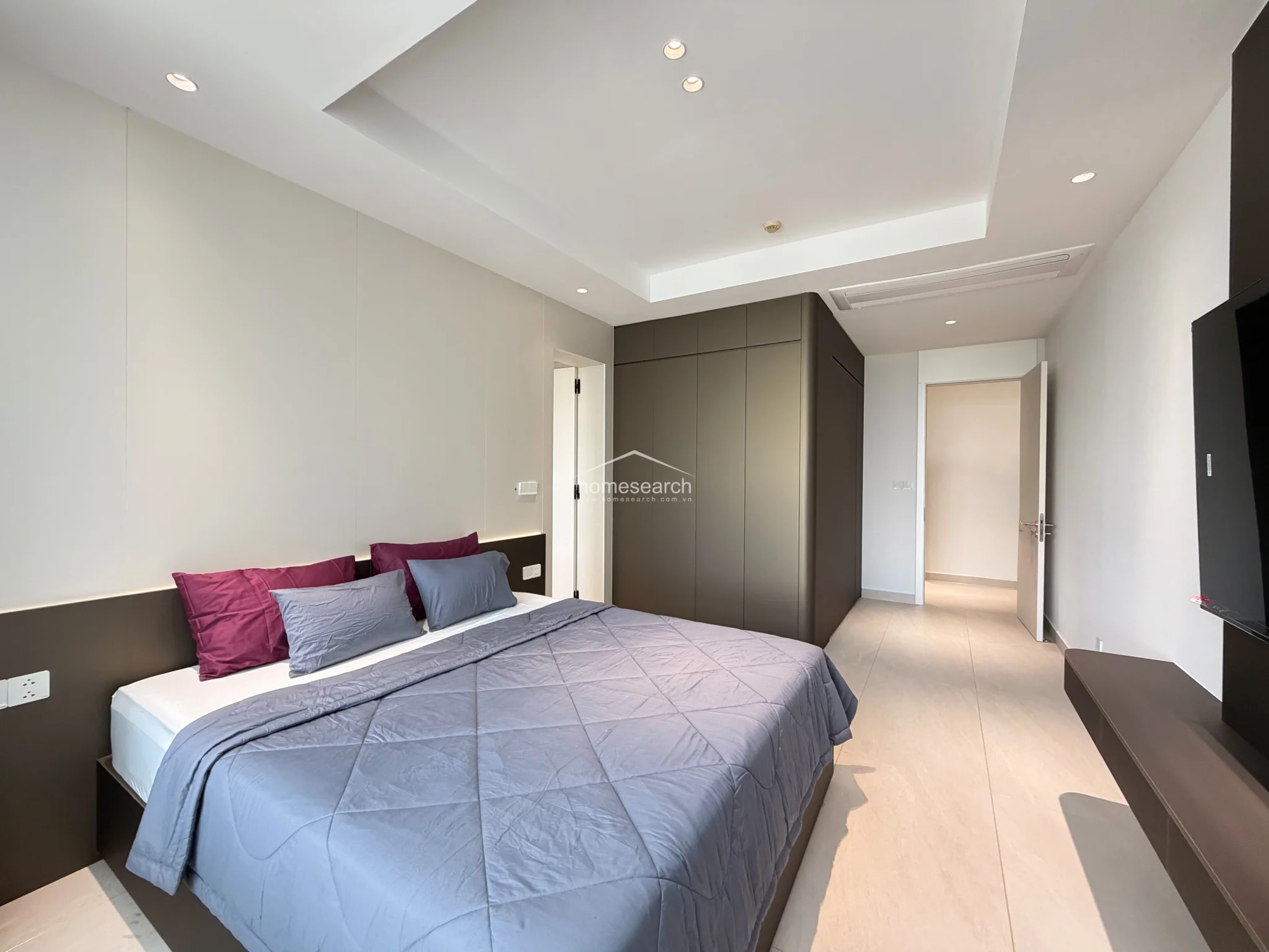 This view highlights the seamless flow of the bedroom, showing the transition from the sleeping area toward the entryway and the built-in wardrobe. The room maintains a consistent color palette of soft greys and deep browns, accented by recessed ceiling lighting that enhances the modern architectural design.