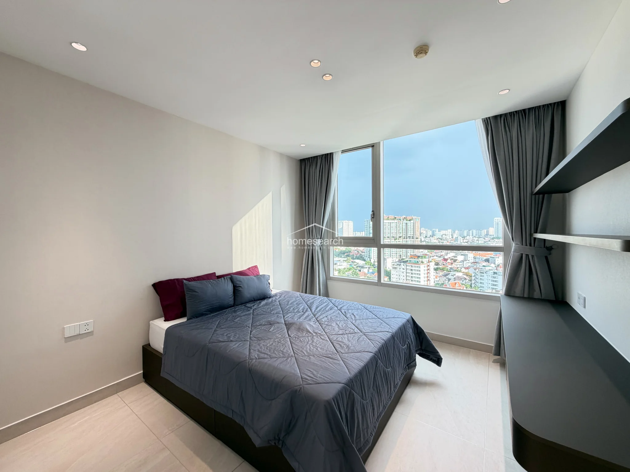 This modern bedroom features a large, floor-to-ceiling window that offers an expansive urban view and fills the space with abundant natural light. The room is finished with light-colored tile flooring and neutral walls, complemented by a sleek, built-in black shelving unit and a matching desk.