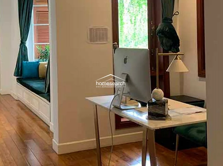 This home office or study area is characterized by warm wood flooring and a built-in window bench seat with plush cushions, offering a cozy nook for reading or relaxing. A modern desk setup with an Apple computer is positioned near a window, creating a bright and functional workspace.