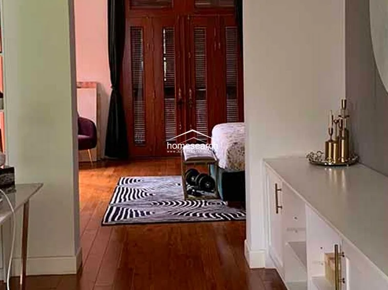 This bedroom or lounge area features rich hardwood flooring and elegant dark curtains that frame a set of traditional wooden louvered doors. The room includes a built-in white storage cabinet with gold hardware, providing a clean contrast to the warm wood tones and adding a touch of refined organization to the space.