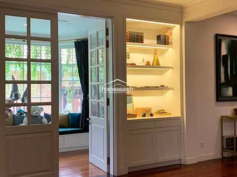 This living space features an elegant transition through French doors into a cozy bay-window nook, which is lined with built-in bench seating and lush garden views. The room is finished with warm hardwood flooring and includes a sophisticated built-in shelving unit with integrated display lighting.