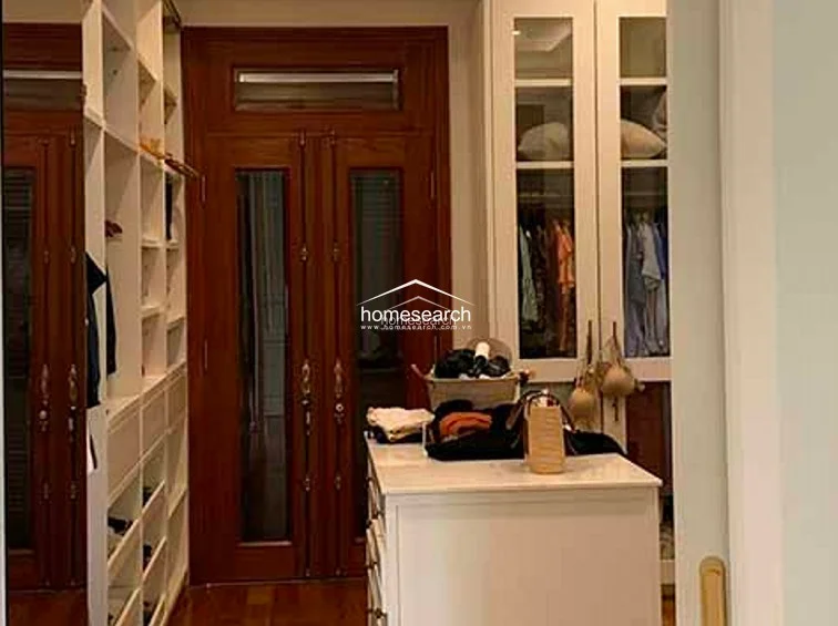 This walk-in closet is meticulously organized with extensive custom white cabinetry, including open shelving and hanging sections behind glass-paneled doors. The room maintains a high-end aesthetic with rich hardwood floors and a central island unit that provides additional storage and a surface for accessories.