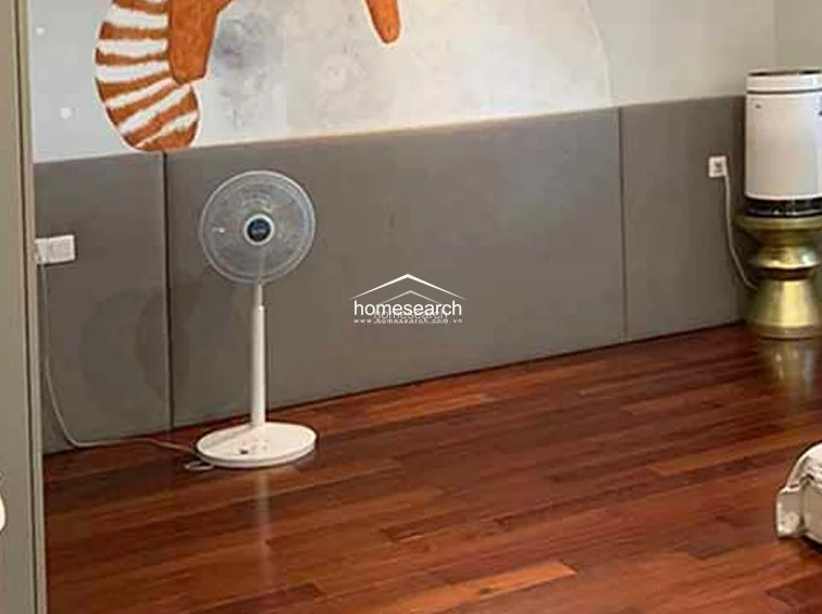 This bedroom area showcases a contemporary design with a plush, grey upholstered headboard wall and whimsical wall art, set against warm-toned hardwood flooring. The room is equipped with a portable electric fan and an air purifier, maintaining a minimalist and well-maintained atmosphere.