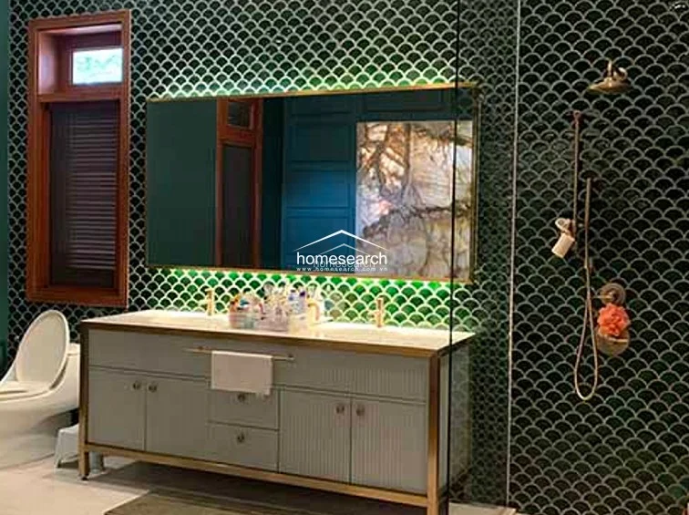 This bathroom showcases a striking design with dark green, scalloped-pattern wall tiles and a large, illuminated mirror above a modern vanity. The gold-toned fixtures and warm lighting create a luxurious, high-end feel, complemented by a clean white countertop and integrated storage cabinets.