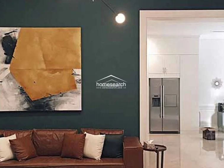 This living area showcases a bold design aesthetic with deep teal walls contrasted by a large, warm-toned abstract art piece and a plush leather sofa. The space flows openly into a bright, neutral-toned kitchen area, highlighting a clean and modern architectural layout.