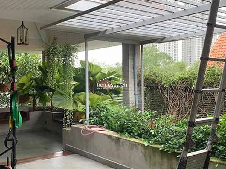 This outdoor terrace space is covered by a slatted pergola, providing a shaded environment for the surrounding potted plants and hanging greenery. The area includes a concrete planter and a metal ladder, creating a functional and serene garden retreat with a view of neighboring architecture.