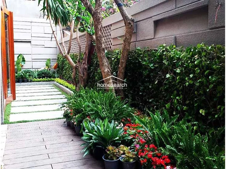 This tranquil garden area features a wooden deck pathway bordered by a stone-tiled walkway and dense, vibrant landscaping. The space is enclosed by a textured concrete privacy wall, creating a secluded and well-maintained outdoor retreat.