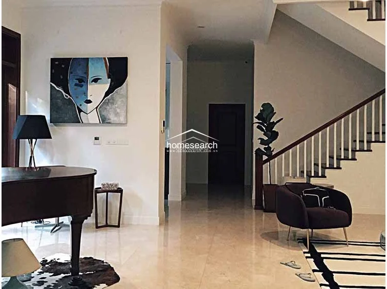 This elegant living area features polished marble flooring and a grand staircase with a wooden handrail and white balusters. The space is tastefully furnished with a piano, a modern armchair, and contemporary wall art, reflecting a sophisticated and well-maintained interior.