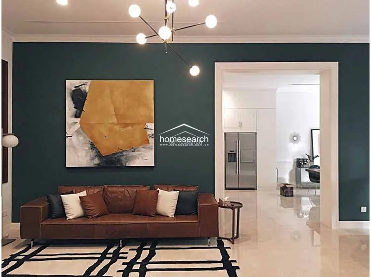 This elegant living room is defined by a deep teal accent wall, a modern leather sofa, and a striking geometric chandelier. The space features polished light-colored flooring and flows directly into an adjacent kitchen area through a wide, open doorway.