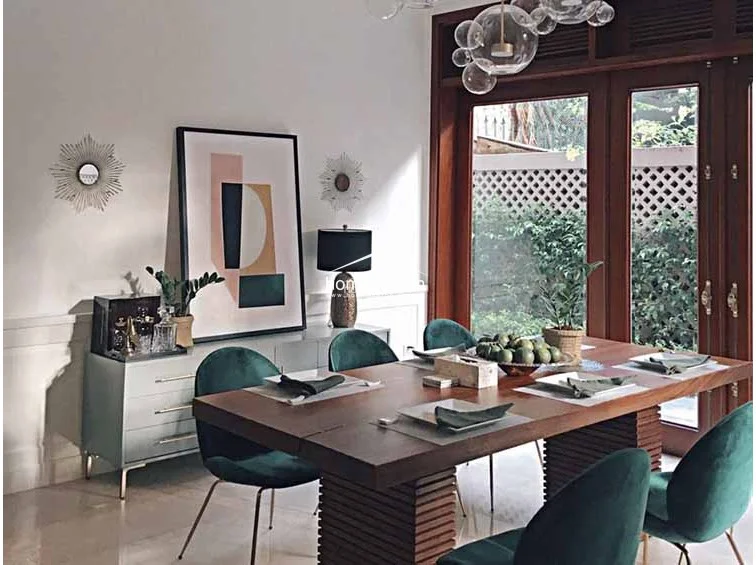 This formal dining area is anchored by a large wooden table with velvet-upholstered chairs and a unique bubble-style light fixture. Large wooden-framed glass doors provide direct access to the outdoor garden area, filling the room with natural light and greenery.