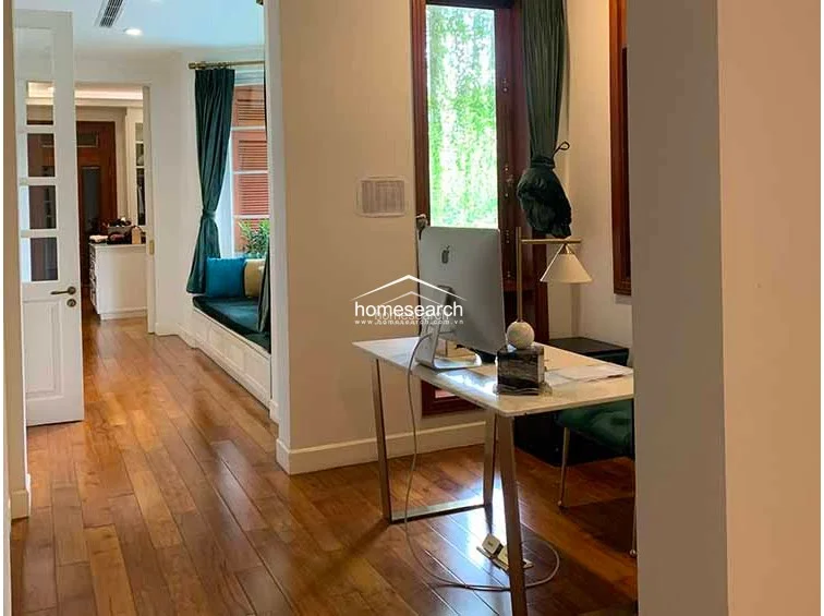 This home office space features warm wood flooring and a built-in window seat with plush cushions, creating a cozy reading or relaxation nook. The room is well-lit by natural light from the large windows and connects seamlessly to the rest of the home through a white paneled door.