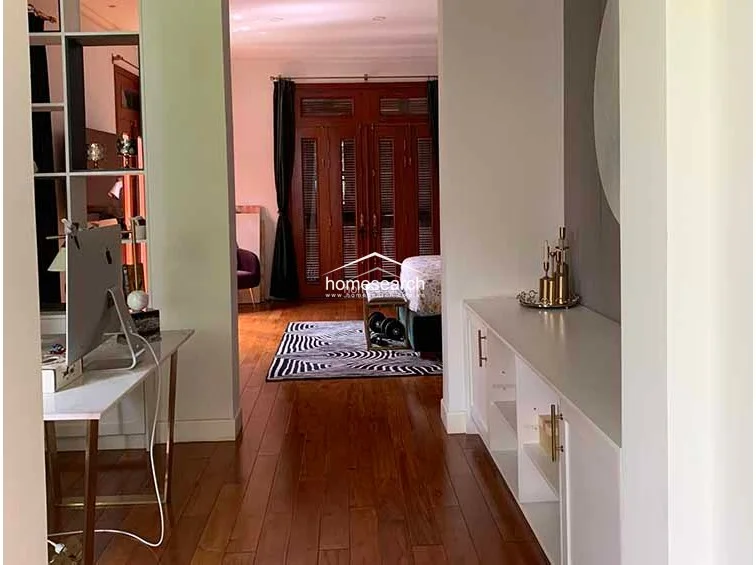 This view captures a transitional hallway or study area featuring rich hardwood flooring and clean white walls. The space is well-organized with a modern desk setup on the left and built-in white cabinetry on the right, leading directly into a bedroom with classic wooden louvered doors.