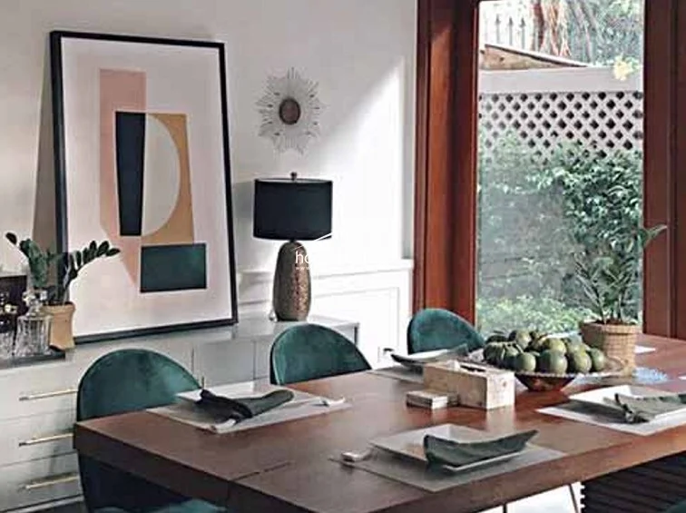 This dining area exudes a sophisticated, warm atmosphere with a wooden table, velvet teal chairs, and elegant decor including a large abstract print and a textured table lamp. Natural light filters in through the large wooden-framed window, which offers a view of a lush green outdoor space.