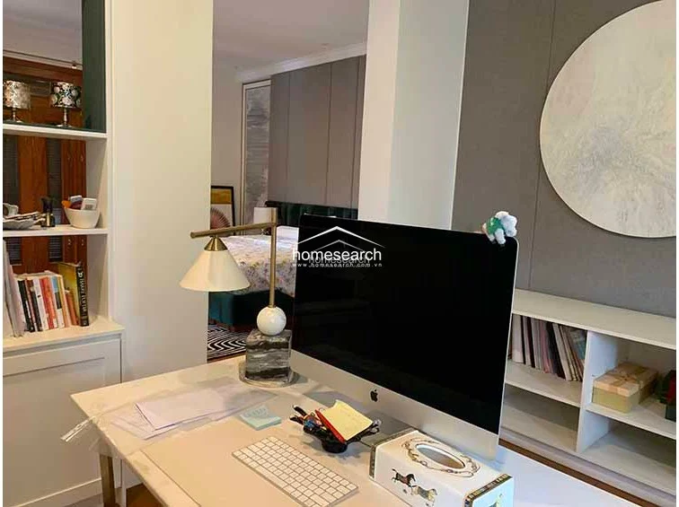 This close-up of the workspace highlights a clean, minimalist aesthetic with a white desk, a modern computer setup, and a stylish brass-accented lamp. The adjacent built-in shelving provides functional storage, while the neutral wall finish and warm wood flooring maintain a cohesive, high-end feel.