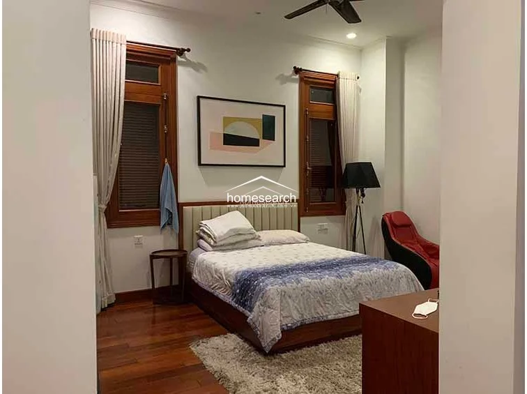 This bedroom features a comfortable bed with neutral bedding, flanked by two large wooden-framed windows that provide ample natural light and a traditional architectural touch. The room is accented by a plush rug, a red lounge chair, and a ceiling fan, all set against a backdrop of warm hardwood floors and white walls.