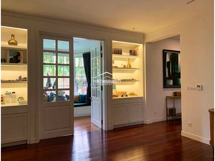 This elegant living area features rich dark hardwood flooring and custom floor-to-ceiling built-in cabinetry with integrated lighting. A set of classic French doors leads into a cozy, sunlit reading nook or window seat area, creating a seamless flow between spaces.