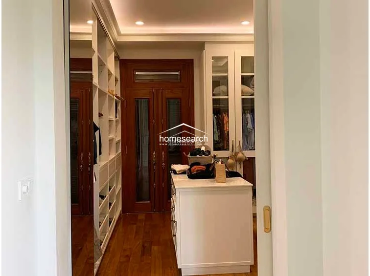 This walk-in closet is highly organized with extensive white cabinetry, glass-fronted wardrobe doors, and a central storage island. The space is finished with warm wood flooring and recessed ceiling lighting, maintaining a clean and upscale aesthetic.