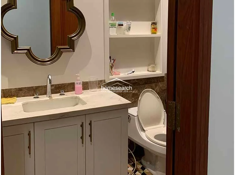 This bathroom features a sophisticated vanity with a marble countertop, a unique quatrefoil-shaped mirror, and built-in recessed shelving for storage. The space is well-maintained, utilizing a combination of neutral tones and decorative floor tiles to create a polished, functional design.