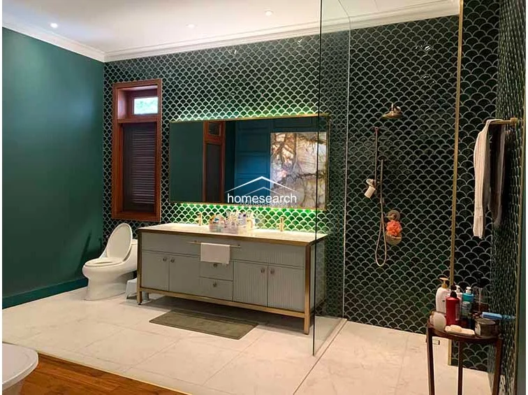 This luxurious bathroom showcases a sophisticated design with deep green scalloped wall tiles and a striking backlit onyx feature wall behind a freestanding clawfoot tub. The space combines rich wood flooring with high-end brass fixtures and a custom vanity, creating an opulent and well-maintained spa-like environment.